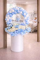 Decorative floral arrangement with balloons and 'Welcome Baby' text in a hospital setting.
